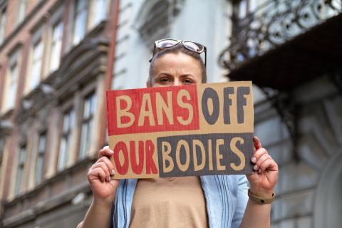 A woman holding a hand-made sign that reads "Ban off out Bodies" in front of city buildings.