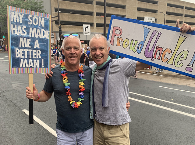 Joe Strauss and Bill Baldus stand smiling, arms around each other and holding two signs. One reads "My gay son has made me a better man!" and the other reads "Proud Uncle!"
