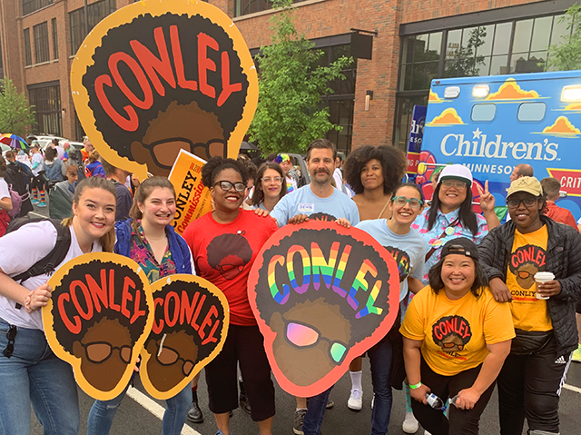 A crowd of Angela Conley supporters and the County Commissioner herself stand with shirts and signs featuring her name and logo, all grinning towards the camera.