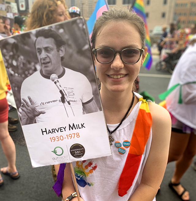 Parker Janssen holds a picket sign featuring Harvey Milk speaking into a microphone which reads "Harvey Milk, 1930-1978" and features the OutFront MN and Stonewall 50th anniversary logos.