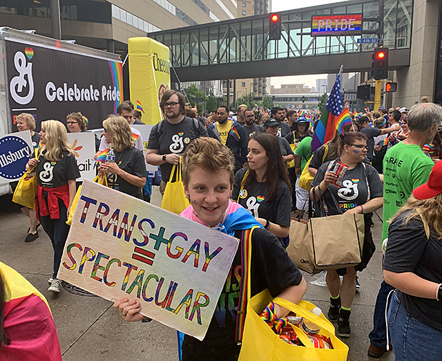 A crowd marches along the parade route in t-shirts featuring the General Mills logo. At the front, one individual holds a sign that reads "Trans + Gay = Spectacular."