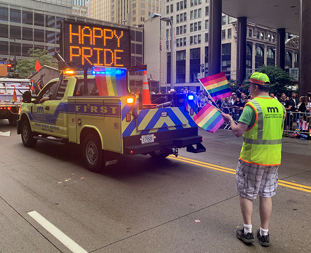 A Minnesota Department of Transportation with a rainbow flag hanging off its back and a digital sign reading "Happy Pride" drives off, with one staff member following behind and holding two smaller rainbow flags.